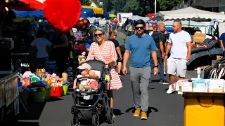 Ein Paar mit einem Baby im Kinderwagen geht eine belebte Straße auf einem Markt entlang, mit einem riesigen roten Herzdekoration über ihnen.