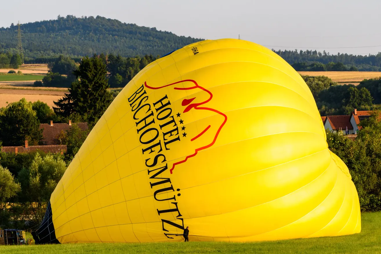 Ein gelber Heißluftballon mit der Aufschrift Hotel Bischoffsmütz ist teilweise aufgeblasen auf einer Grasfläche. Dahinter steht eine Person in der Nähe des Korbes. In der Ferne sind Bäume und ein Hügel zu sehen.