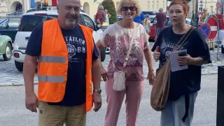 Drei Personen stehen vor einem historischen Gebäude mit roten Dächern und lächeln für ein Foto. Ein Mann in einer orangefarbenen Weste steht links, eine Frau mit Sonnenbrille und rosa Hosen steht in der Mitte und eine weitere Frau in einem schwarzen Shirt steht rechts.