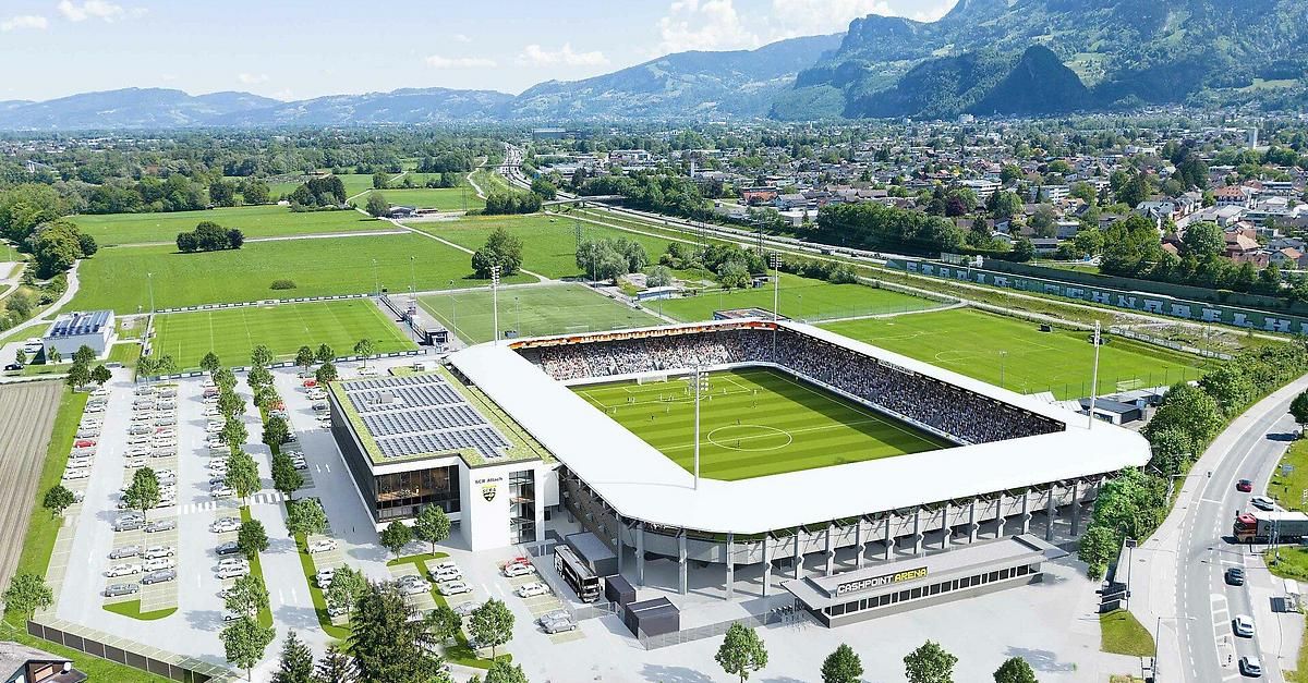 Eine Luftaufnahme eines großen Fußballstadions mit grünen Feldern und einer Bergkette im Hintergrund.