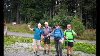 Vier Männer stehen zusammen auf einem Kiesweg, lächeln und geben Daumen hoch. Sie sind fürs Wandern gekleidet und halten Wanderstöcke. Der Hintergrund zeigt dichten grünen Wald.