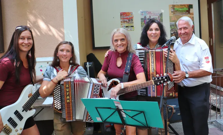Drei Frauen spielen Musikinstrumente, darunter Akkordeon, Gitarre und ein weiteres Akkordeon, lächeln und posieren für ein Foto. Sie stehen vor einem Mikrofon. Hinter ihnen ist eine Wand mit Plakaten.