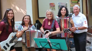 Drei Frauen spielen Musikinstrumente, darunter Akkordeon, Gitarre und ein weiteres Akkordeon, lächeln und posieren für ein Foto. Sie stehen vor einem Mikrofon. Hinter ihnen ist eine Wand mit Plakaten.