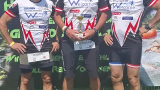 Drei Männer, wahrscheinlich Triathleten, stehen auf einem Grasfeld. Sie tragen Radtrikots mit dem Wort 'Willkommen' und halten eine Trophäe. Der Mann auf der linken Seite hat einen Namensschild mit dem Namen Daniel. Der Mann in der Mitte trägt eine Sonnenbrille. Hinter ihnen steht ein Banner mit dem Text 'Olympisch' und einigen Logos.