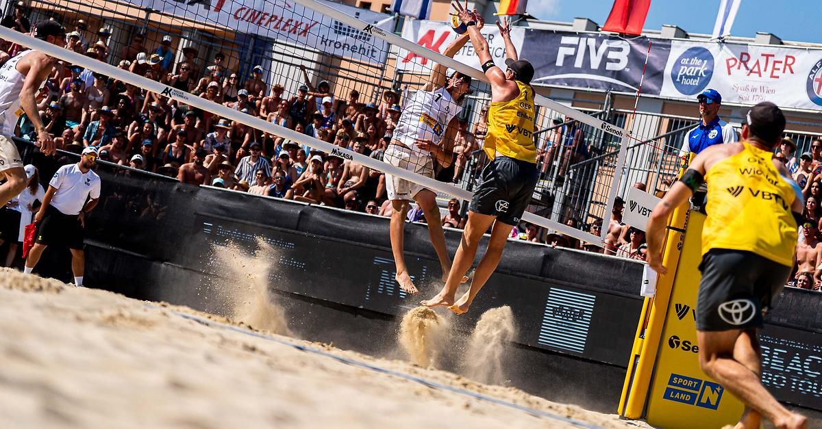 Zwei Männer spielen Beachvolleyball, einer blockt den Ball am Netz, während der andere springt. Viele Zuschauer sehen von den Tribünen zu.