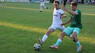 Ein Fußballspiel auf einem Rasenfeld, Spieler in weißen und grünen Trikots, ein Spieler in Weiß bereitet den Ball zum Schuss vor, ein anderer in Grün verteidigt.