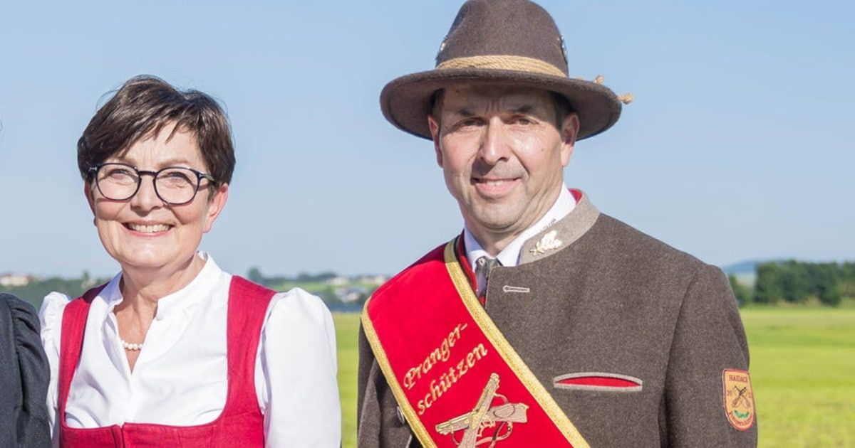Ein Mann und eine Frau stehen zusammen auf einem Feld und lächeln. Der Mann ist in einer traditionellen Uniform mit einem roten Schal und einem Hut gekleidet. Die Frau trägt eine rote Schürze.