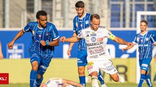 Zwei Fußballspieler spielen ein Match, einer in Blau und einer in Weiß, wobei einer versucht, den anderen zu tackeln. Dahinter beobachtet ein weiterer Spieler in Blau.