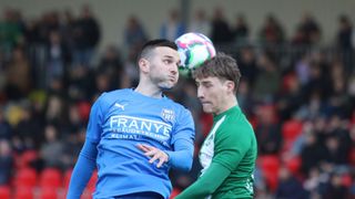 Zwei Fußballspieler in blauen und grünen Trikots kämpfen um den Ball, während eine Menge im Hintergrund zusieht.