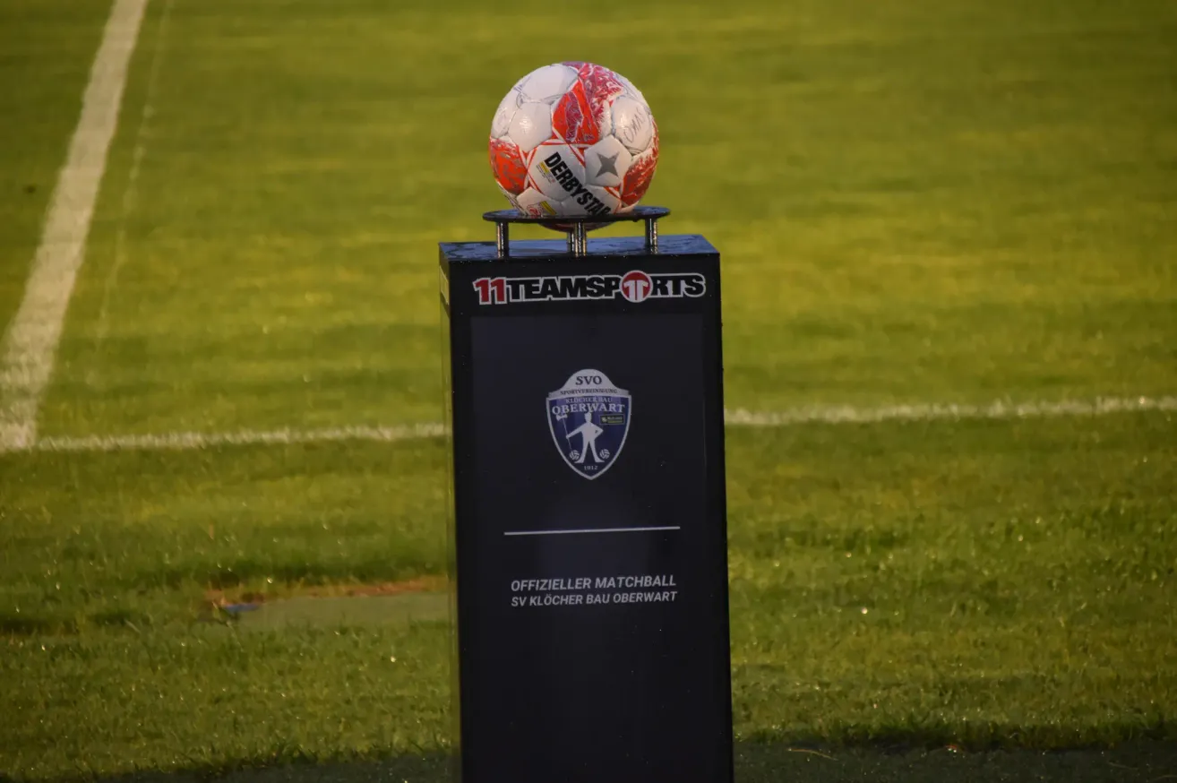 Ein Fußball auf einem Ständer mit dem Logo von SV Oberwart, auf dem die Texte 'Offizieller Matchball' und 'SV Klocher Bau Oberwart' zu sehen sind. Der Ball steht auf einem Rasenfeld.