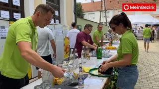 Eine Gruppe von Menschen in grünen Shirts bereitet Getränke an einem Tisch im Freien mit Flaschen, Gläsern und einem gelben Tuch zu. Einige tragen Brillen, und einer trägt eine Armbanduhr. Ein Kühlschrank ist im Hintergrund.