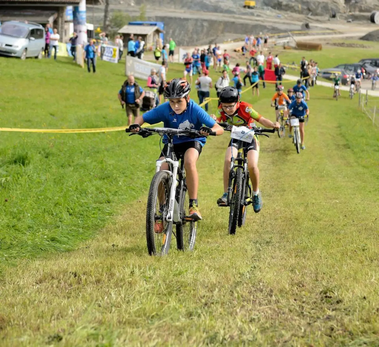 Eine Gruppe von Kindern fährt auf einem Rasenhügel mit ihren Fahrrädern, während eine Menge Menschen von der Seite zusieht. Die Fahrer tragen Helme und Radsporttrikots. Der Rasen ist üppig, und im Hintergrund befinden sich eine Mauer und geparkte Autos.