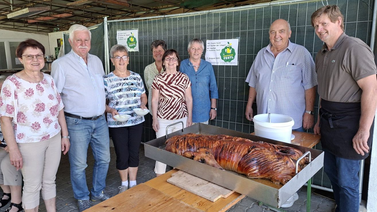 Eine Gruppe von Menschen steht hinter einem Tisch mit einem großen gebratenen Schwein darauf. Sie scheinen bei einer Veranstaltung zu sein, mit Schildern am Zaun hinter ihnen.