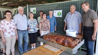 Eine Gruppe von Menschen steht hinter einem Tisch mit einem großen gebratenen Schwein darauf. Sie scheinen bei einer Veranstaltung zu sein, mit Schildern am Zaun hinter ihnen.