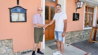 Zwei Männer stehen vor einem Gebäude. Sie schütteln sich die Hände. Beide lächeln. Hinter ihnen befindet sich eine Tür und ein Schild.