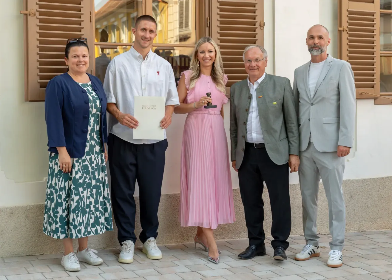 Fünf Personen stehen vor einem Gebäude. Ein Mann hält ein Zertifikat und eine Auszeichnung, während eine Frau neben ihm ein rosa Kleid trägt. Sie lächeln. Hinter ihnen stehen zwei weitere Männer und eine Frau, alle in formeller Kleidung. Die Umgebung scheint ein formeller Anlass zu sein.