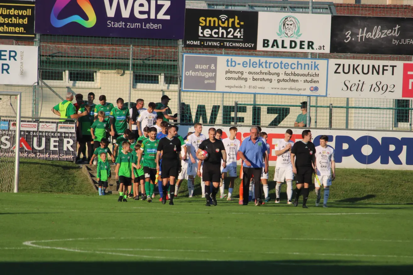 Fußballspieler in grünen und weißen Trikots, begleitet von Schiedsrichtern, gehen über ein grünes Feld. Im Hintergrund befinden sich Werbebanner für Weiz, smart-tech24 und Balder.at.