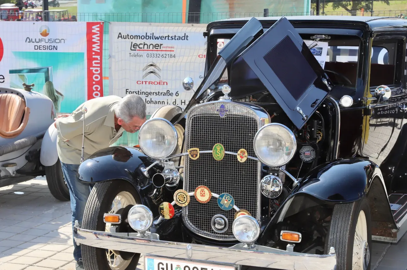 Ein Mann untersucht die offene Motorhaube eines klassischen Autos, das mit verschiedenen Abzeichen verziert ist und vor einem Banner für Automobilwertstadt Lechner geparkt ist.