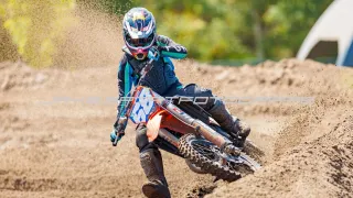 Ein Motocross-Fahrer mit Helm und Schutzausrüstung fährt auf einer Erdbahn und erzeugt eine Staubwolke. Der Fahrer befindet sich in einer leicht geneigten Position, wobei sich das Motorrad nach rechts neigt. Im Hintergrund sind Bäume und ein klarer Himmel zu sehen.