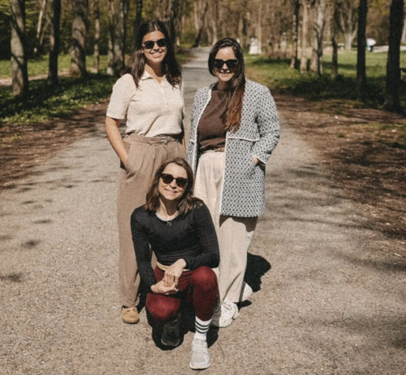Drei Frauen, alle mit Sonnenbrillen, posieren auf einem Kiesweg umgeben von Bäumen. Eine kniet, eine steht zwischen zwei anderen.