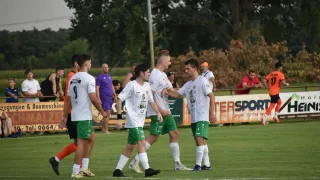 Fußballspieler in weißen und grünen Trikots gratulieren sich gegenseitig auf einem Feld mit einem Banner im Hintergrund.