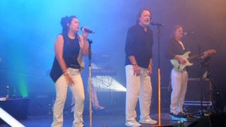 Eine Frau singt in ein Mikrofon, während ein Mann hinter ihr steht. Sie sind auf einer Bühne mit blauer Beleuchtung und einem Keyboarder im Hintergrund.