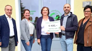 Drei Menschen stehen lächelnd vor einem Gebäude. Eine Frau hält ein Schild mit der Aufschrift 'Lasberger Bauerinnen, €400,- Spende vom Pflanzenmarkt.'