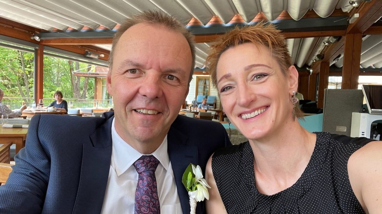 Ein Mann und eine Frau lächeln auf einem Foto. Sie haben beide kurze Haare. Der Mann trägt eine Krawatte mit Paisleymuster. Die Frau hat eine Blumenbrosche auf der Schulter. Sie stehen vor einem unscharfen Hintergrund mit einem Gebäude und Bäumen.