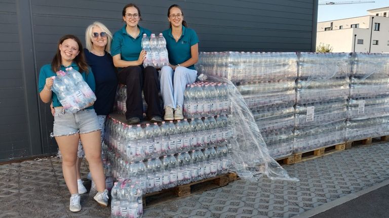Vier Frauen stehen neben einem großen Stapel Plastikflaschen, zwei von ihnen halten eine Packung Flaschen. Sie scheinen für ein Foto zu posieren.