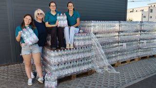 Vier Frauen stehen neben einem großen Stapel Plastikflaschen, zwei von ihnen halten eine Packung Flaschen. Sie scheinen für ein Foto zu posieren.