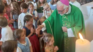 Ein Bischof in einer grünen Robe segnet Kinder in einer Kirche, einige mit geflochtenen Haaren, die traditionelle Kleidung tragen.