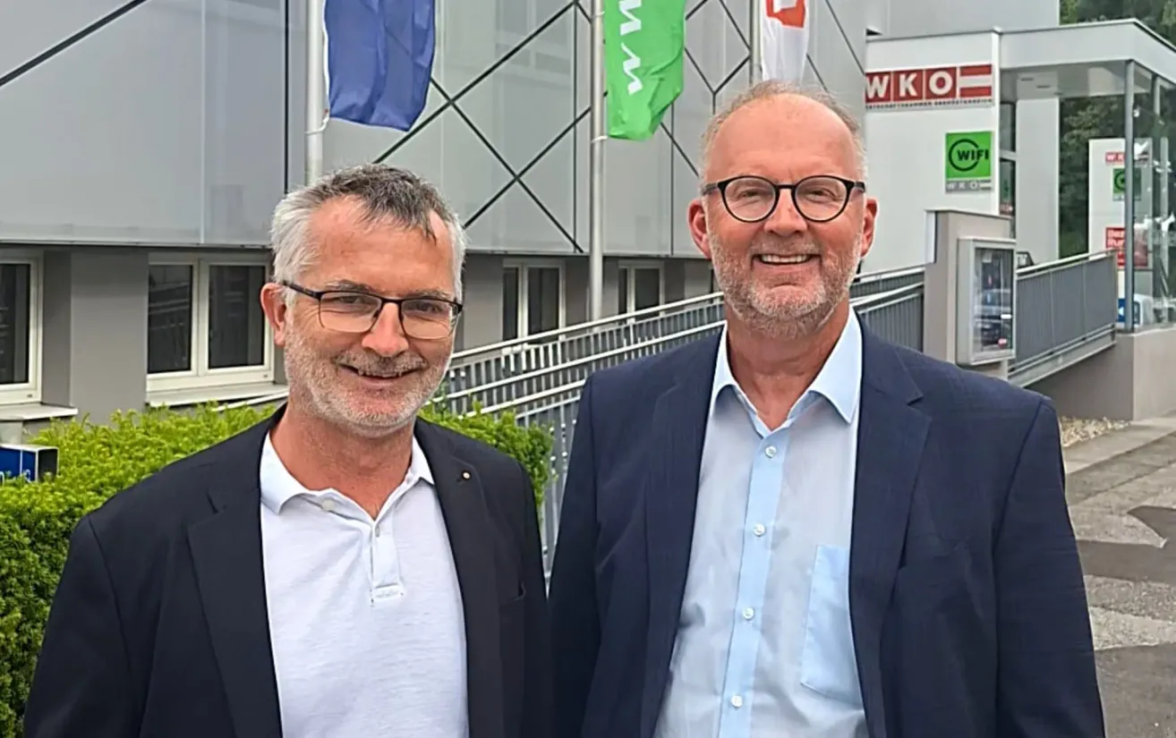 Zwei Männer, beide mit Brille und Business-Anzug, stehen vor einem Gebäude mit Fahnen im Hintergrund.