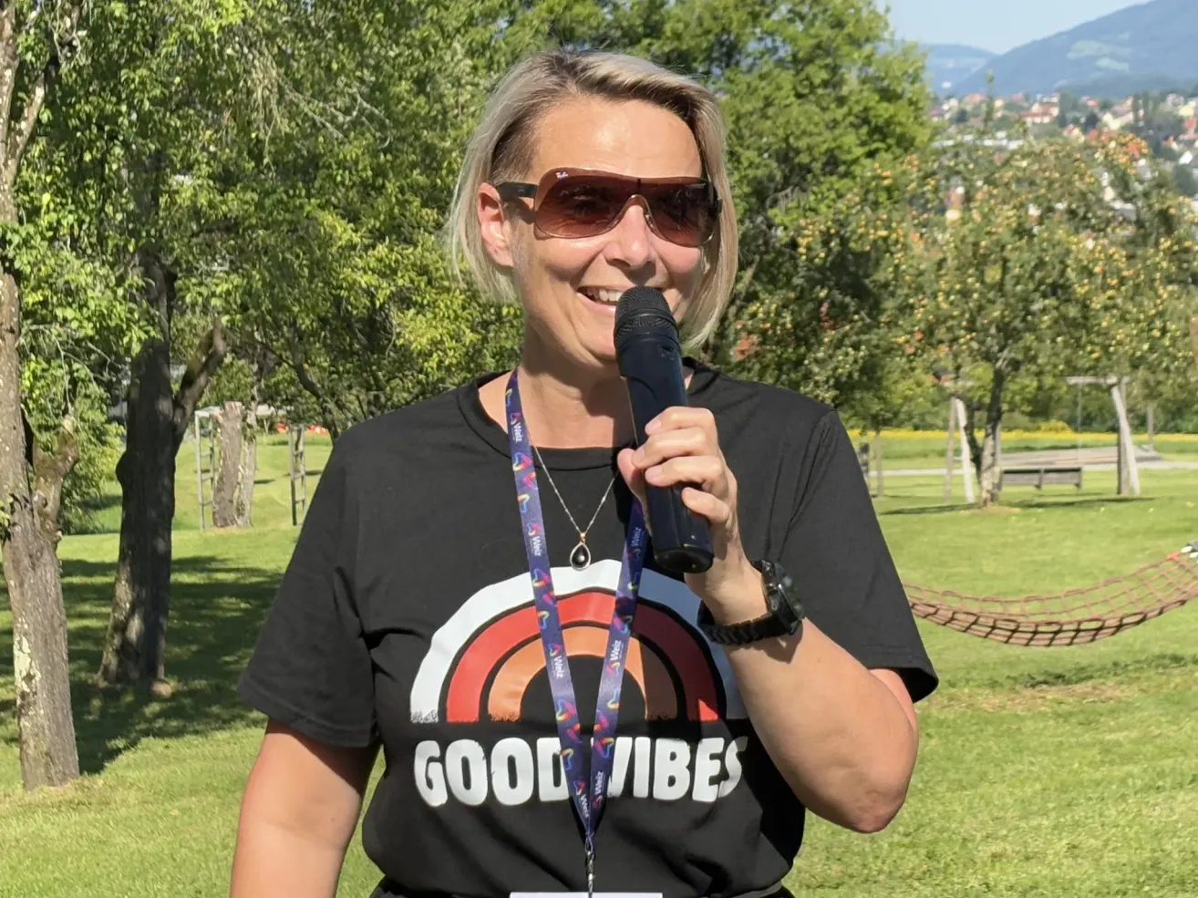 Eine Frau in einem schwarzen T-Shirt mit den Worten 'Good Vibes' steht in einem Park, hält ein Mikrofon und lächelt. Sie trägt eine Sonnenbrille und einen Ausweis. Der Hintergrund zeigt grünen Rasen, Bäume und in der Ferne Berge.