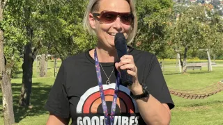 Eine Frau in einem schwarzen T-Shirt mit den Worten 'Good Vibes' steht in einem Park, hält ein Mikrofon und lächelt. Sie trägt eine Sonnenbrille und einen Ausweis. Der Hintergrund zeigt grünen Rasen, Bäume und in der Ferne Berge.