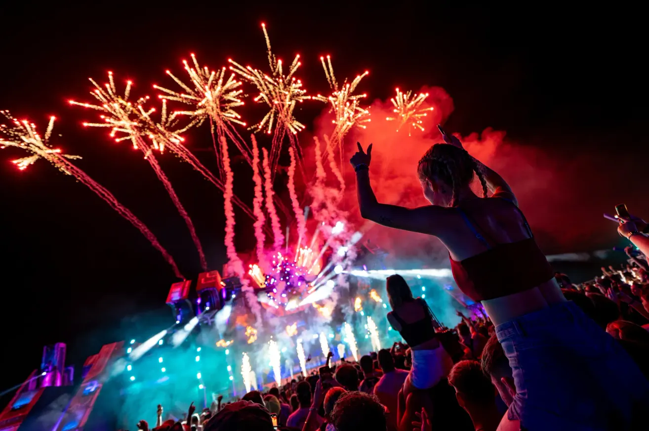 Ein lebhaftes Nachtmusikfestival zeigt eine große Menschenmenge, eine Frau, die ihre Hände hebt, und ein spektakuläres Feuerwerk.