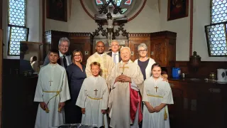 Eine Gruppe von Menschen versammelt sich in einer Kirche, lächelt und posiert für ein Foto. Sie tragen religiöse Kleidung. Der Priester steht mit gefalteten Händen in der Mitte. Hinter ihnen befindet sich ein Altar mit einem Kruzifix und einem Buntglasfenster.