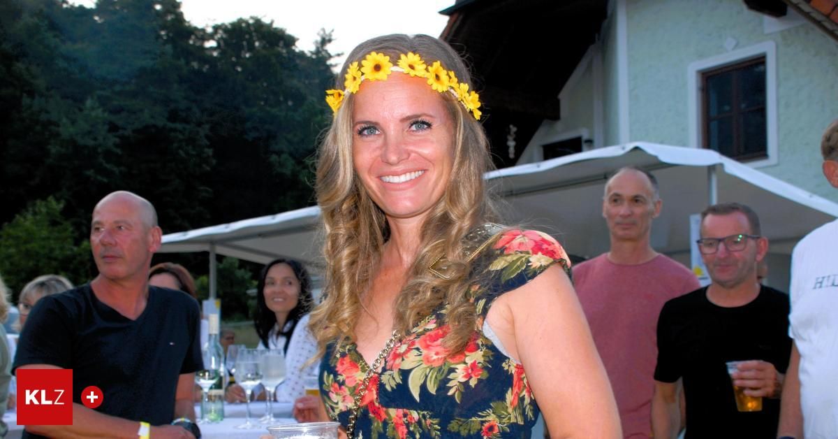 Eine Frau mit einem Sonnenblumen-Kopfband und einem Blumenkleid steht lächelnd bei einer Outdoor-Veranstaltung. Hinter ihr sitzen Leute an einem Tisch, und ein Mann in einem rosa T-Shirt steht neben einem Gebäude.