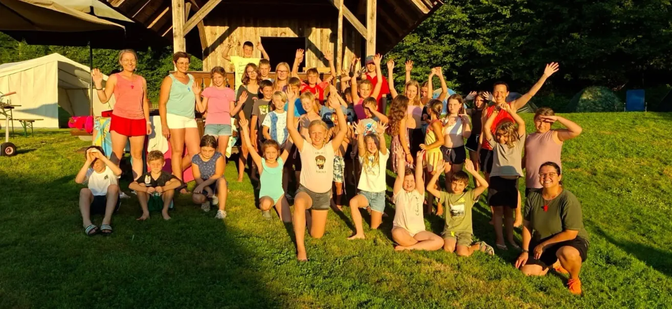 Eine Gruppe von Kindern und Jugendlichen posiert vor einer Holzhütte, einige knien, andere stehen mit erhobenen Händen, an einem sonnigen Tag.