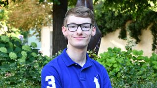 Ein Mann in einem blauen Poloshirt mit der Nummer 3, lächelnd und mit Brille. Im Hintergrund befinden sich Grünpflanzen und ein Baum.