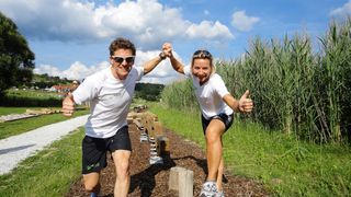 Zwei Menschen in weißen Shirts und schwarzen Shorts posieren mit erhobenen Daumen, tragen Sonnenbrillen und Turnschuhe auf einem Rasenfeld mit Holzpfosten und blauem Himmel.