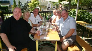 Eine Gruppe von fünf Personen, einschließlich eines Priesters, sitzt an einem Tisch im Freien. Sie haben Gläser mit Wein und Flaschen auf dem Tisch, mit einem Gebäude und Bäumen im Hintergrund.