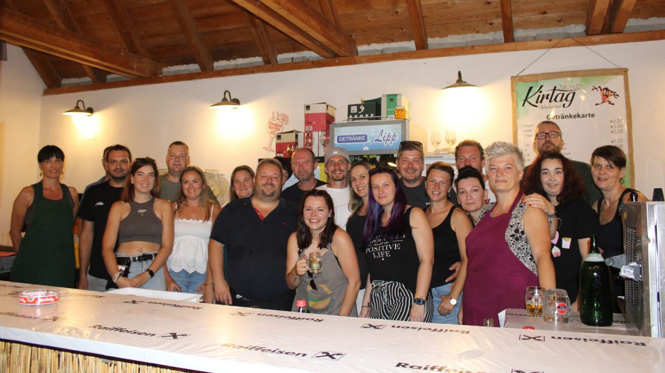 Eine Gruppe von Menschen posiert für ein Foto in einem Raum mit Holzbalken, einer weißen Wand und einem Tisch. Eine Frau in der Vorderfront hält ein Getränk. Ein Kühlschrank mit dem Wort Lipp steht an der Wand. Ein paar Lampen sind an der Decke montiert.