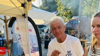 Ein älterer Mann steht unter einem Zelt, lächelt und trägt ein weißes T-Shirt. Im Hintergrund gehen Menschen an Schirmen vorbei, an einem Schild, das auf Trinkwasser hinweist.