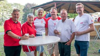 Fünf Menschen stehen lächelnd um einen Tisch mit Getränken. Sie tragen passende rote und weiße T-Shirts. Hinter ihnen steht ein Pavillon und Menschen genießen die Umgebung im Freien.