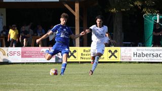 Zwei Fußballspieler in blauen und weißen Trikots kämpfen auf dem Feld, einer von ihnen tritt den Ball. Dahinter stehen mehrere Personen in der Nähe eines Tisches und eines Baumes.