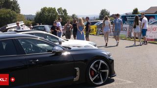 Eine Gruppe von Menschen ist um Autos auf einer Straße versammelt, mit einem Van und geparkten Autos im Hintergrund. Bäume und ein Berg sind in der Ferne sichtbar.