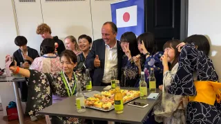 Eine Gruppe von Menschen versammelt sich um einen Tisch mit Essen und Getränken. Ein Mann im Anzug gibt ein Daumen hoch. Hinter ihm macht eine Frau eine Friedensgeste. Sie tragen alle traditionelle japanische Kleidung.