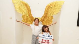 Zwei junge Mädchen posieren für ein Foto vor einem goldenen Engelsflügel-Hintergrund. Sie halten ein Schild mit der Aufschrift 'Basilika Frauenkirchen Engel Fotopoint'.