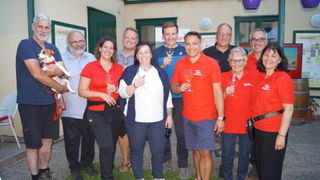 Eine Gruppe von Menschen steht zusammen, hält Weingläser in der Hand und lächelt, vielleicht bei einer Veranstaltung. Sie stehen vor einem Gebäude.