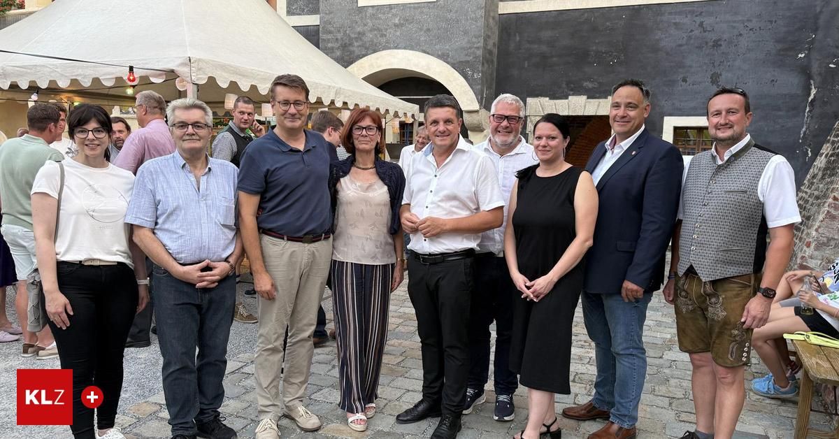 Eine Gruppe von formell gekleideten Menschen posiert für ein Foto vor einem Gebäude mit Zelten und Personen im Hintergrund.